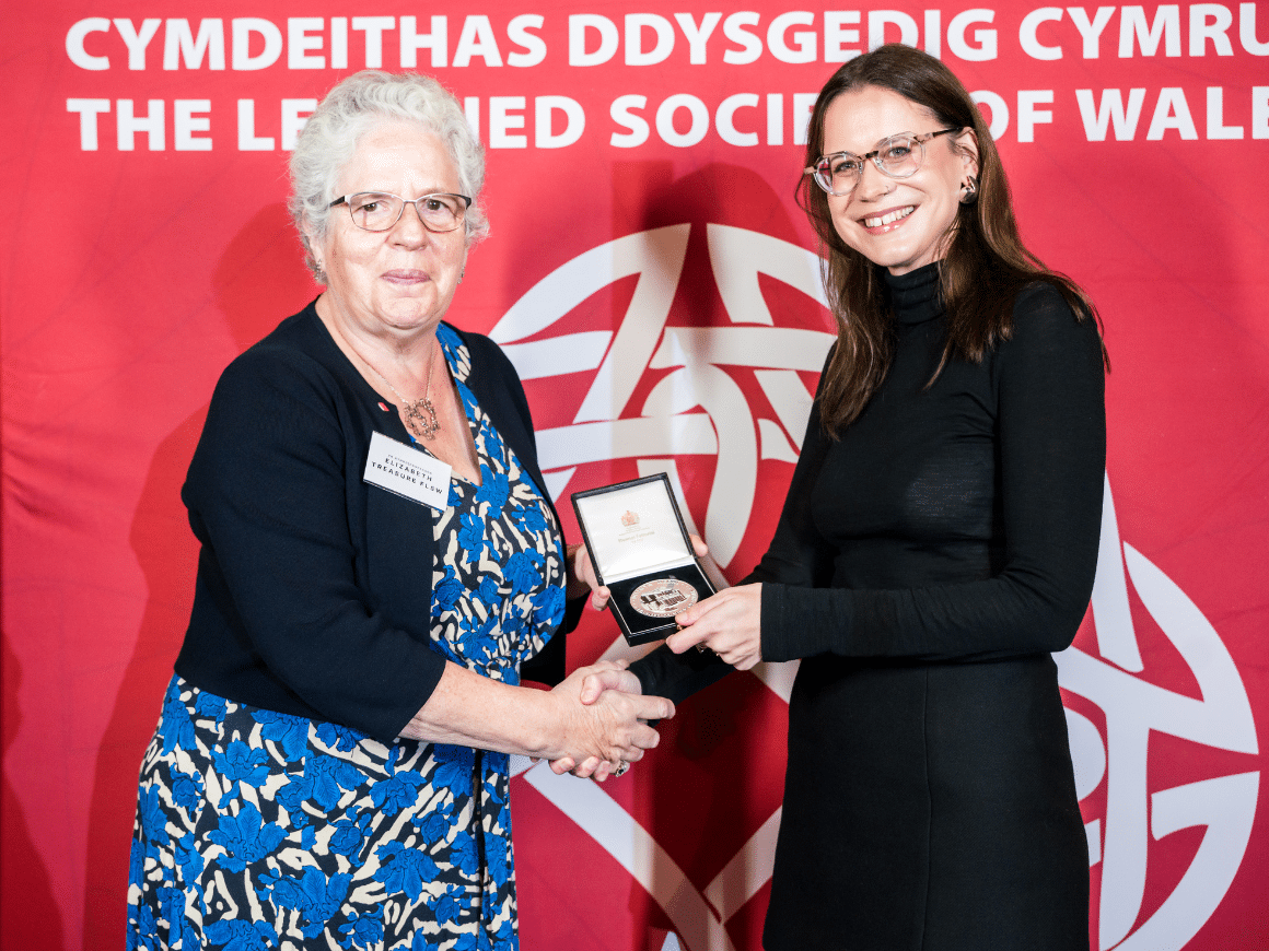 Two women, one holding a Learned Society of Wales medal, shaking hands in fron of an LSW banner.