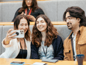 Two women and a man sitting in a lecture hall talking a selfie