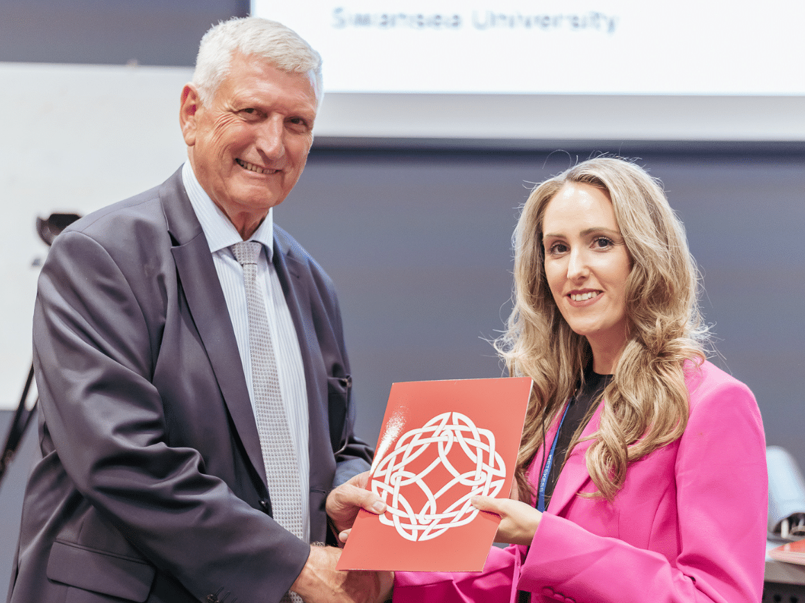 A woman and a man shaking hands while holding a brochure with the Learned Society of Wales logo on it.