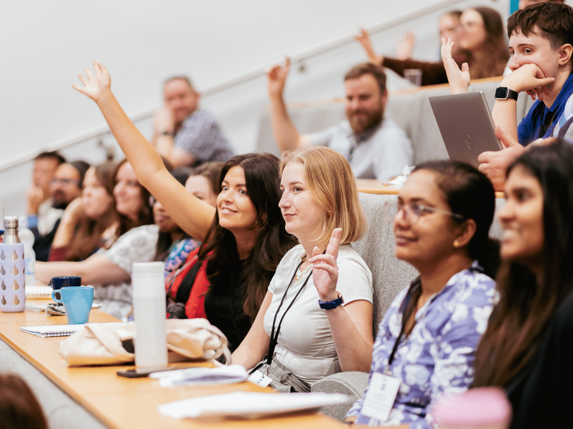An audience sitting in a lecture hall, several of them with their arms raised in the air.