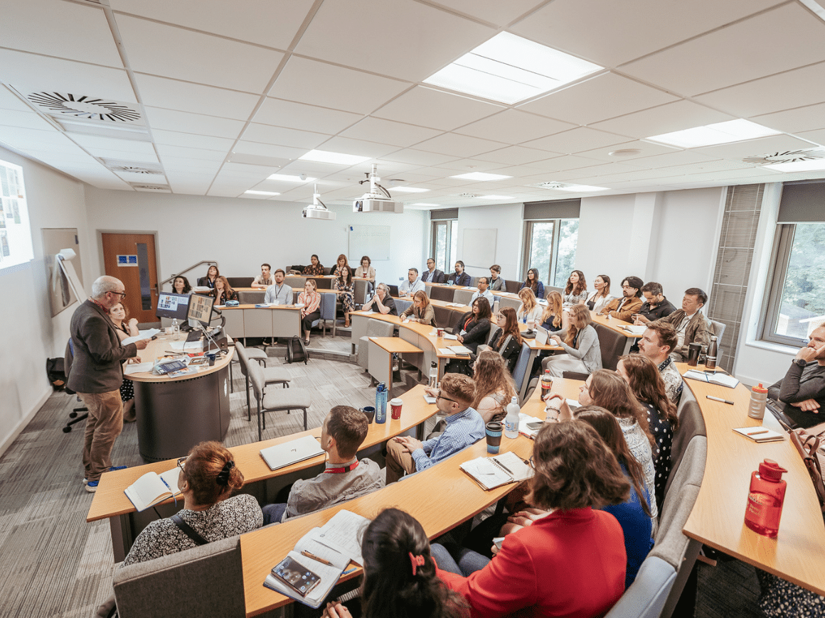 A full seminar room with the audience at semi-circular rows of desks listening to a man speaking.