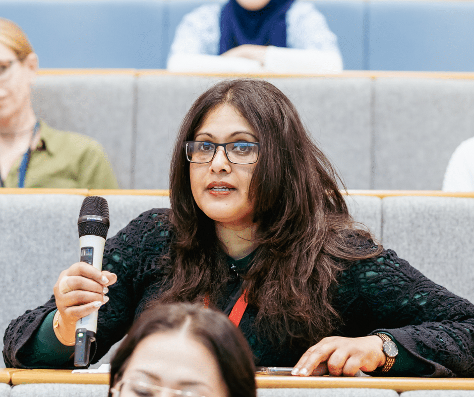 A woman, sitting in a lecture hall, holding a microphone.