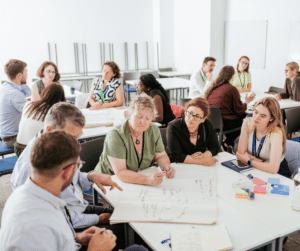 A group of people sit around a table with a large sheet of paper on which they are writing.