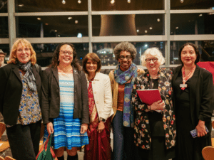 Six women of different ages and ethnicity, all smiling, and standing in a row inside the Welsh Senedd.