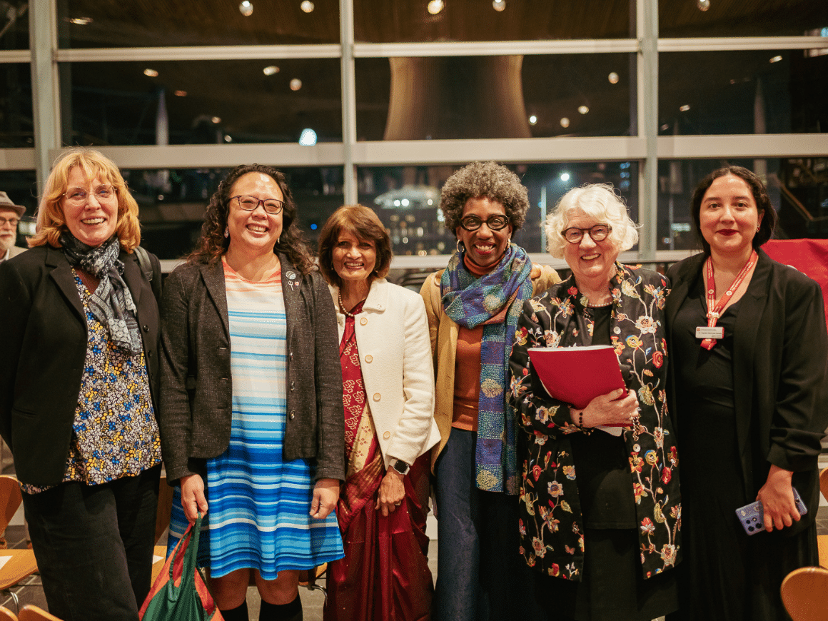 Six women of different ages and ethnicity, all smiling, and standing in a row inside the Welsh Senedd.