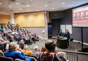 A lecture hall full of people with a man at the front delivering a talk from a lectern.