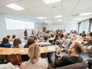 A seminar room with attendees sitting at semi circular desks listening to a presentation.