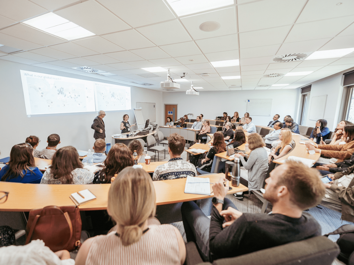 A seminar room with attendees sitting at semi circular desks listening to a presentation.
