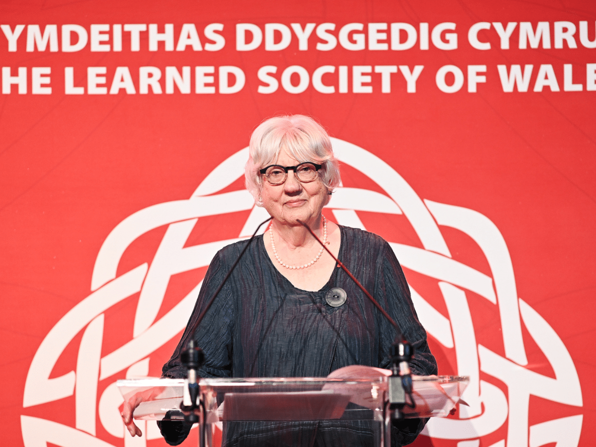 A woman at a lectern in front of a Learned Society of Wales banner stares straight at the camera.
