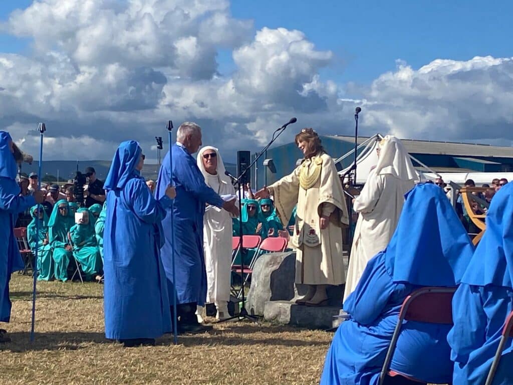 A woman in white robes shaking hands with a man in blue robes in an outdoor venue at the Eisteddfod.