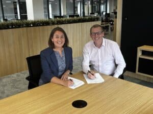 A man and a woman at a table both each signing a form and smiling at the camera.