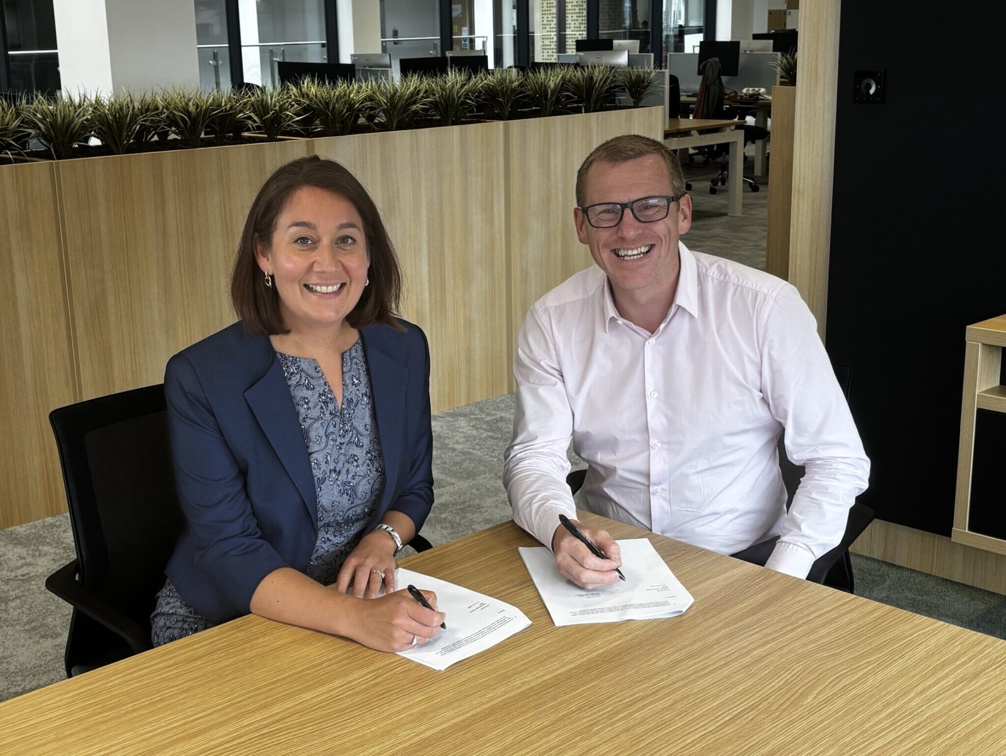 A man and a woman at a table both each signing a form and smiling at the camera.