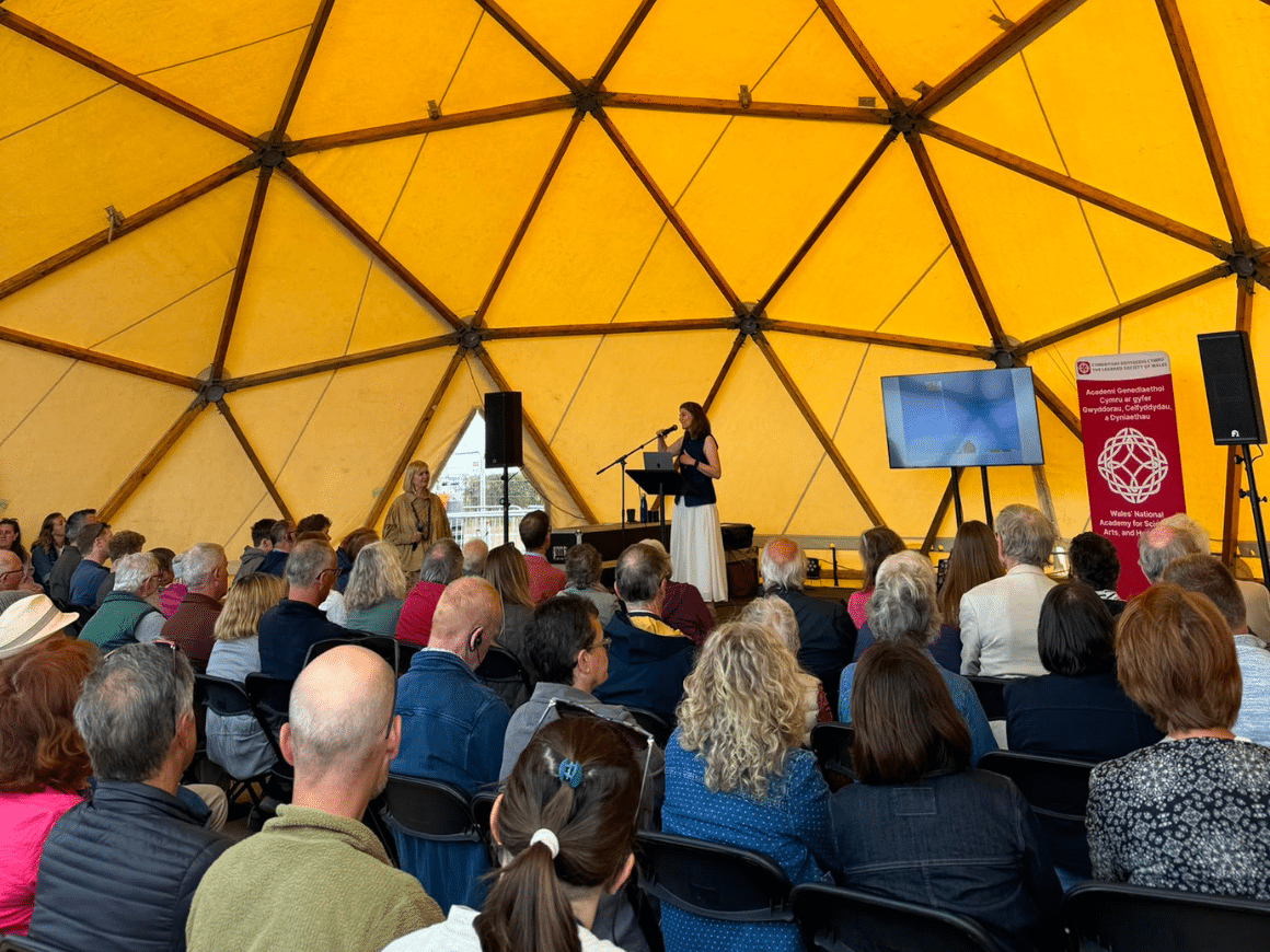 A woman at a lectern delivering a talk to a large crowd in a tent at the Eisteddfod.